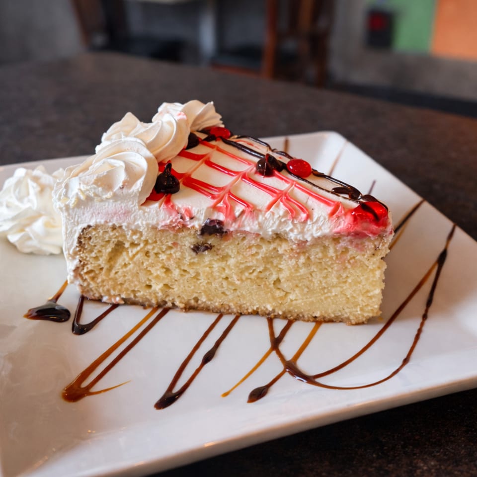 A slice of Tres Leches cake with whipped cream, red and chocolate drizzle, and berries.