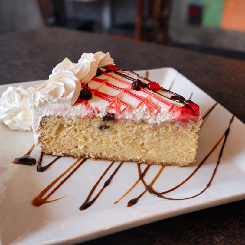 A slice of Tres Leches cake with whipped cream, red and chocolate drizzle, and berries.